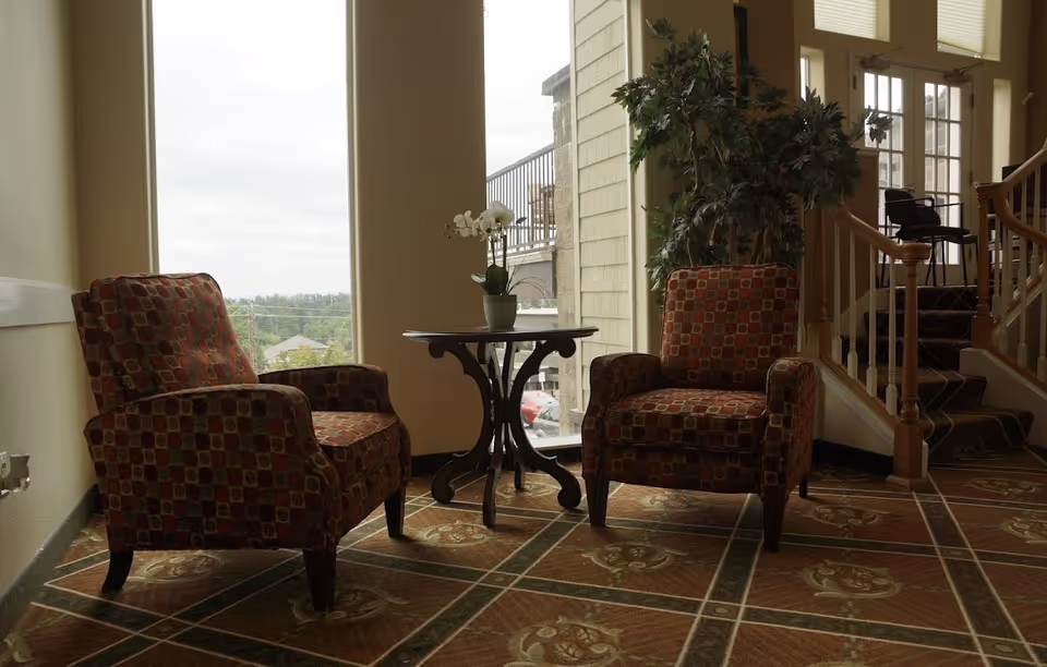 Two patterned armchairs flanking a small round table by large windows in a lounge area with a potted plant and a nearby staircase.
