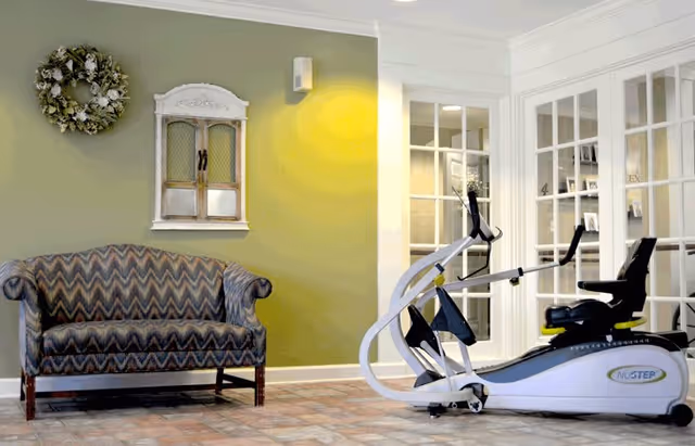 A room with a patterned upholstered loveseat against a green wall decorated with a wreath and a small cabinet. Next to the loveseat is a NuStep exercise machine on a tiled floor. The background shows white-framed glass doors leading to another room.