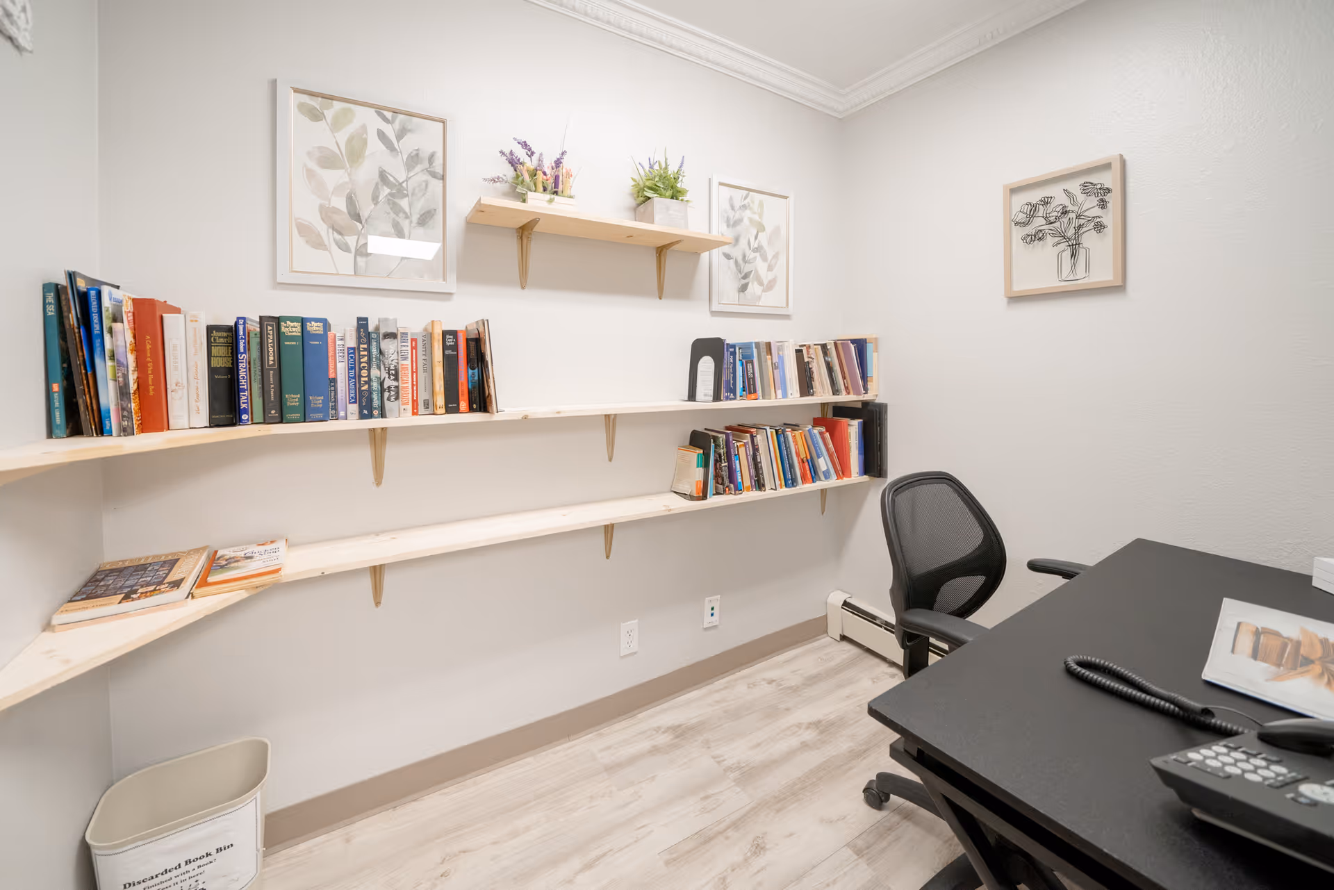 A small office or study room with light-colored walls and wood flooring. There are three wooden shelves mounted on the walls holding various books. Two framed botanical prints and two small potted plants are displayed on the walls and shelves. A black office chair and a black desk with a telephone and a magazine are visible in the corner. A white bin labeled 'Discarded Book Bin' is on the floor.