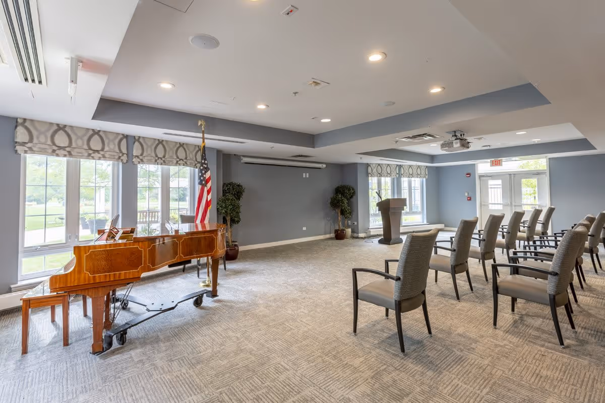 Spacious interior meeting room with a grand piano, American flag, podium and rows of chairs by large windows.