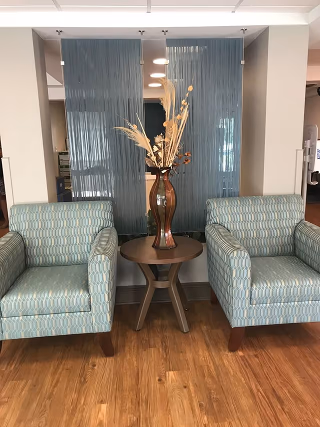 Two patterned armchairs with wooden legs are placed on either side of a small round wooden table holding a tall decorative vase with dried plants. Behind the table and chairs is a blue string curtain hanging from the ceiling, partially obscuring the background. The floor is wooden, and the setting appears to be a calm, indoor seating area.