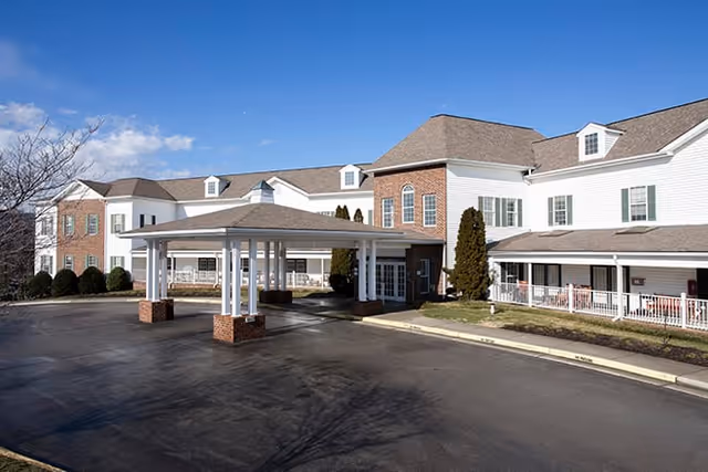 Exterior view of a senior living facility building with white siding and brick accents, featuring a covered entrance with white pillars and a paved driveway under a clear blue sky.