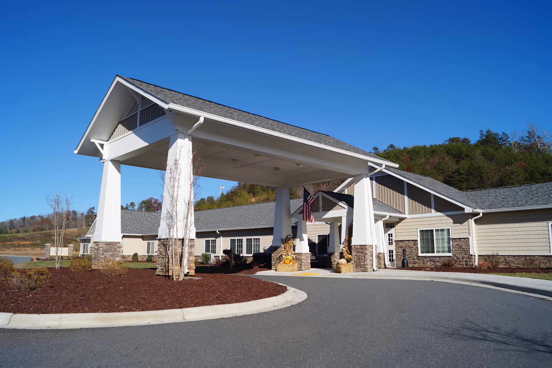 Exterior view of Franklin House facility showing the entrance with a large covered driveway supported by white pillars with stone bases. The building has beige siding with stone accents and a gray shingled roof. There are some small trees and landscaping around the driveway, and an American flag is displayed near the entrance. The sky is clear and blue.