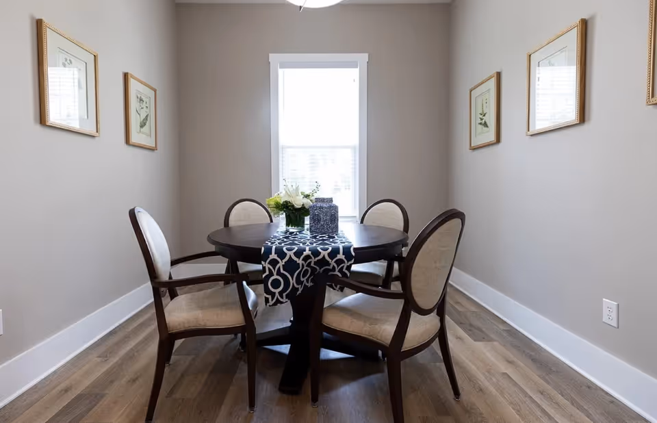Small dining room with a round table, four upholstered chairs, a decorative runner and a window centered on the back wall.