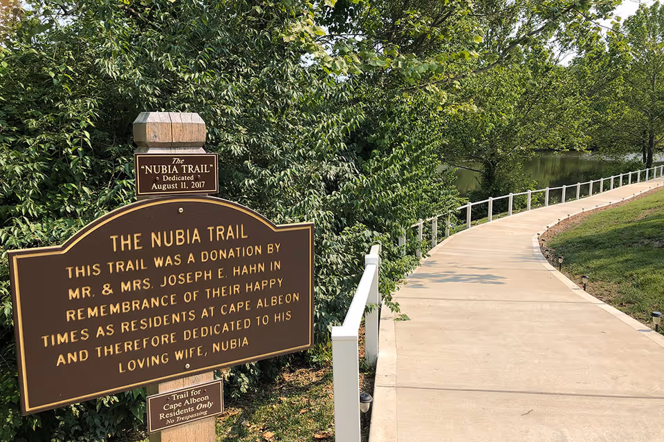 A paved walking trail named The Nubia Trail with a brown sign detailing its dedication and donation by Mr. and Mrs. Joseph E. Hahn in remembrance of their happy times as residents at Cape Albeon. The trail is bordered by a white railing on one side and lush green trees and bushes on the other, with a body of water visible in the background.