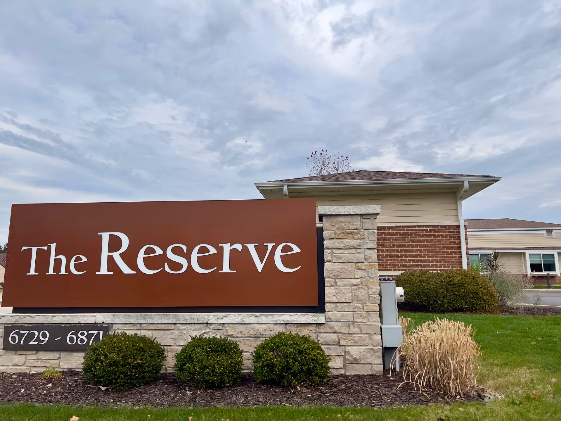 A large stone and brown sign reading "The Reserve" in front of a low brick building and cloudy sky.
