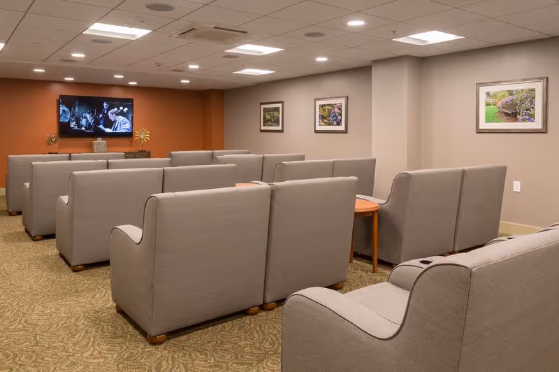 A cozy common area with multiple rows of gray upholstered sofas facing a wall-mounted television. The walls are painted in neutral tones with three framed pictures of flowers and plants. The carpet has a patterned design, and the ceiling has recessed lighting.