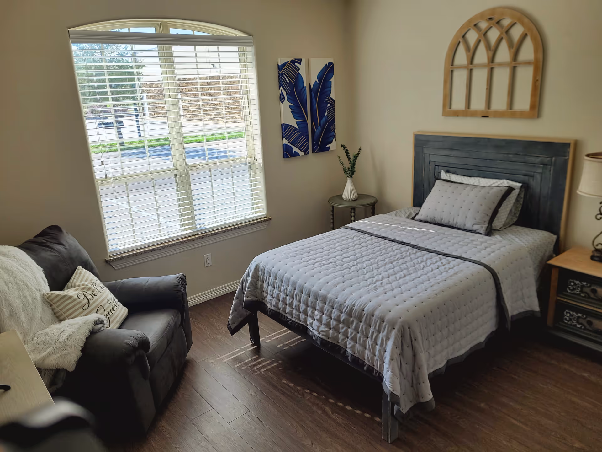 Sunlit bedroom featuring a single bed with gray bedding, an armchair by a large window, and decorative wall art.