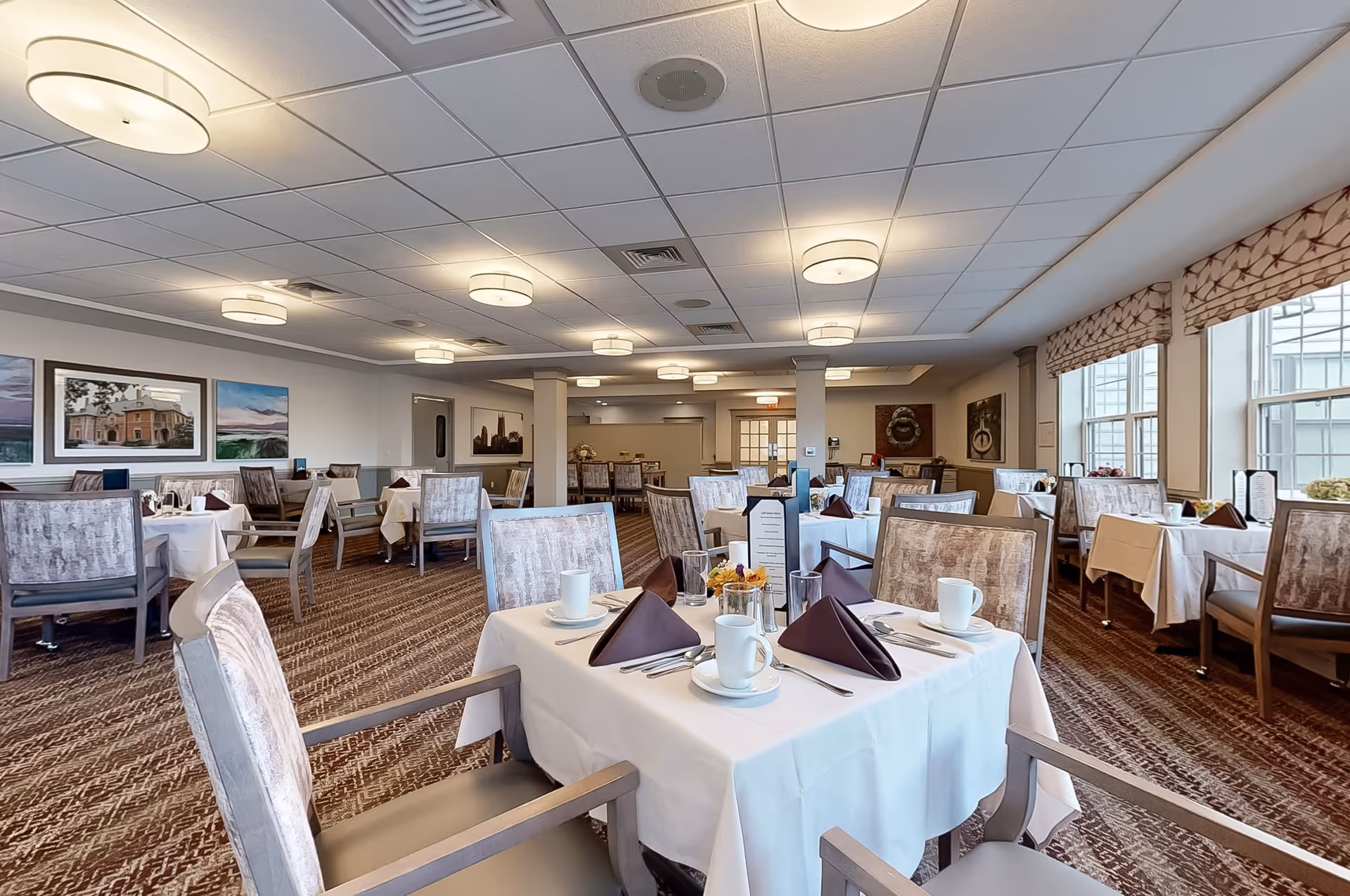 A spacious dining room with multiple tables covered in white tablecloths, each set with white cups, plates, silverware, and folded brown napkins. The room has patterned carpet flooring, cushioned chairs, large windows with patterned valances allowing natural light, and framed artwork on the walls. Ceiling lights provide additional illumination.