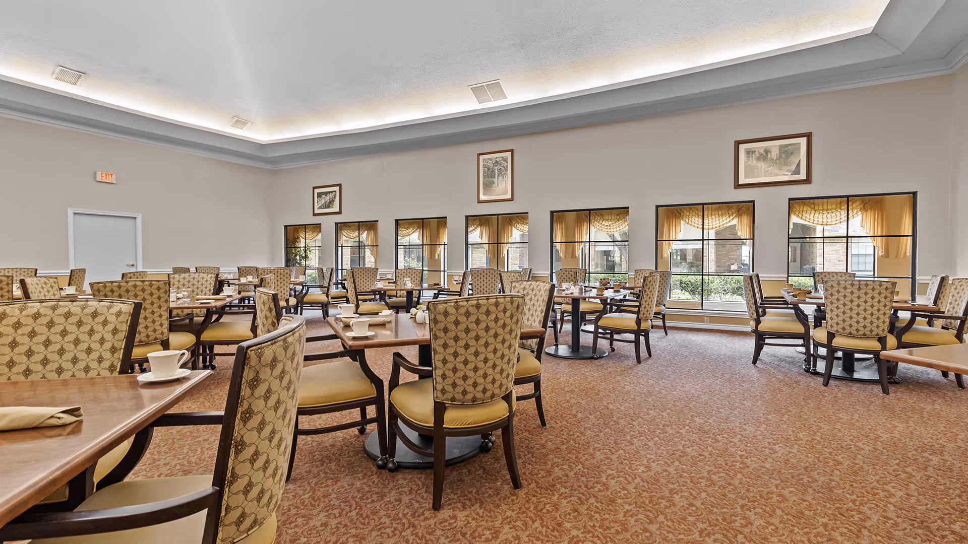 Spacious dining room with multiple tables and patterned upholstered chairs under a recessed ceiling and large windows.
