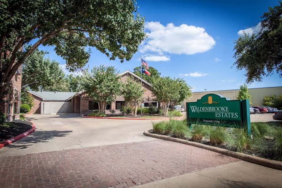 Exterior view of a retirement living facility with a driveway, trees, and a green sign that reads 'Waldenbrooke Estates Retirement Living' under a blue sky with some clouds.