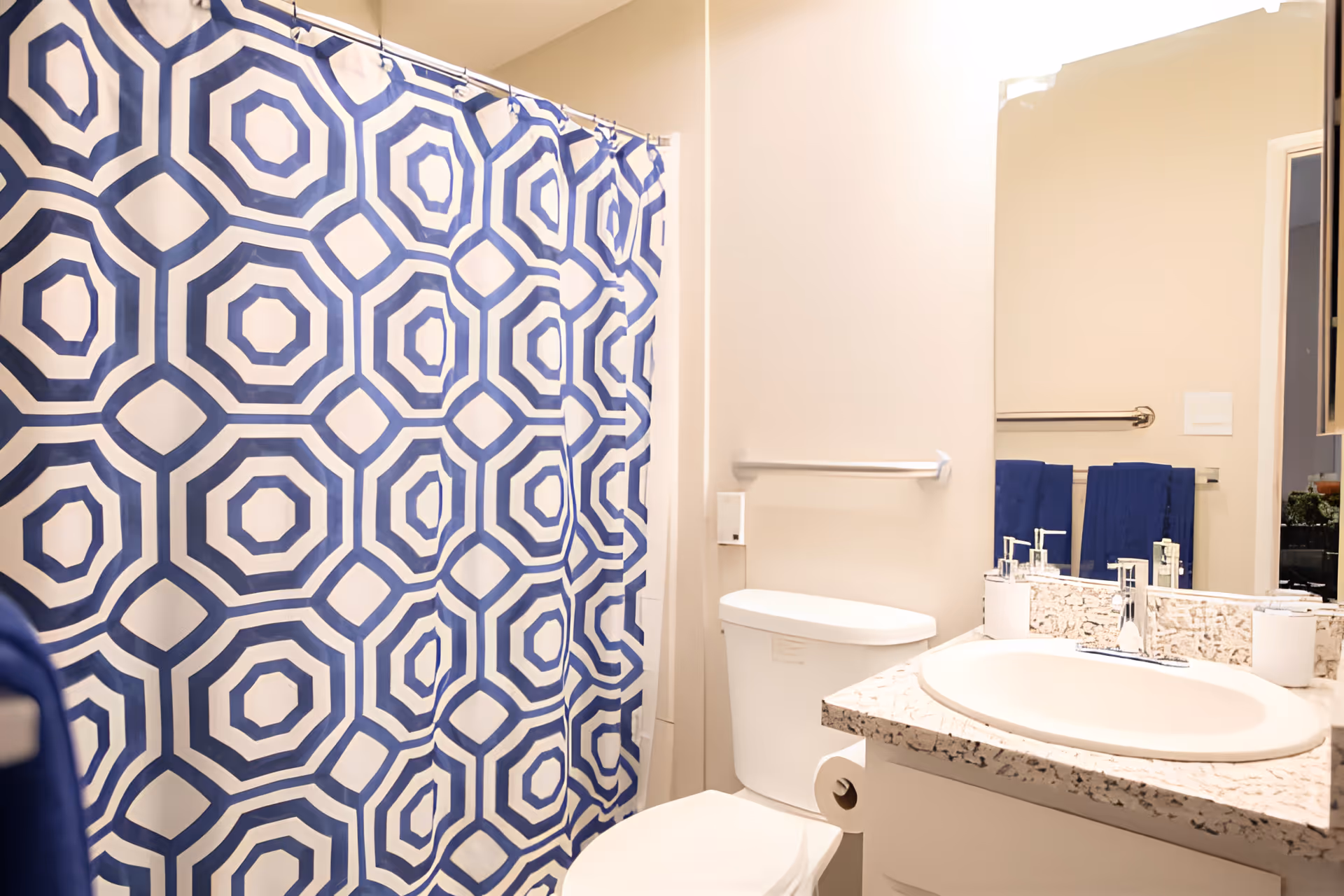 Bright bathroom featuring a blue geometric shower curtain, toilet, sink and mirror with a speckled countertop.