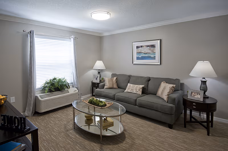 A cozy living room with a gray sofa adorned with three decorative pillows, two side tables each with a lamp and framed photos, a glass oval coffee table with a decorative centerpiece, a window with closed blinds and gray curtains, and a piece of abstract artwork hanging on the wall.