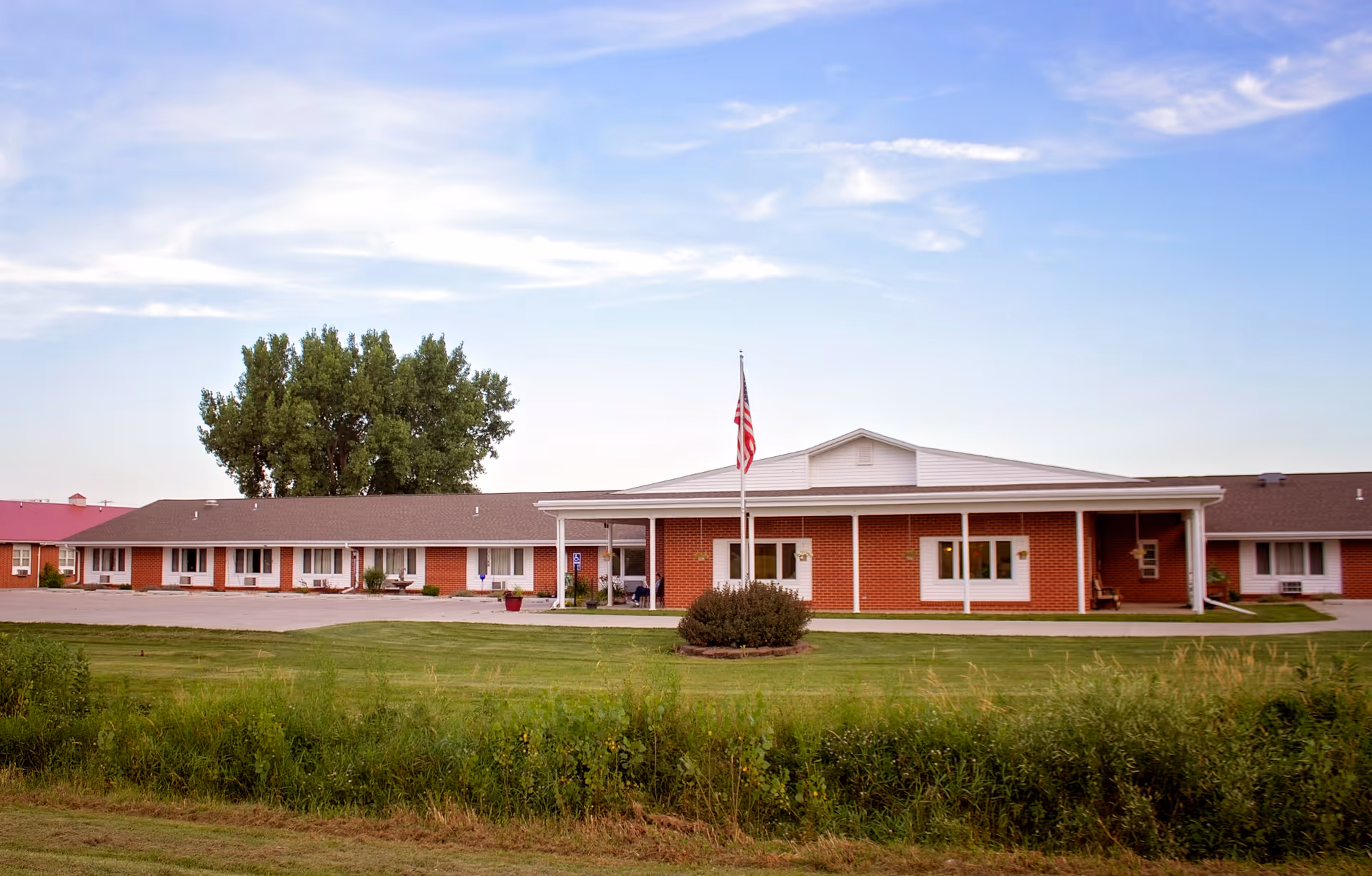 Single-story brick senior living facility front with an American flag on a flagpole and a grassy lawn under a blue sky.