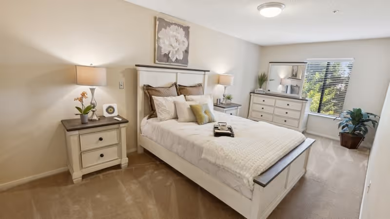 A bright and neatly arranged bedroom featuring a white wooden bed with beige and white bedding, two matching nightstands with lamps and decorative plants, a dresser with a large mirror, a window with blinds letting in natural light, and a potted plant in the corner.