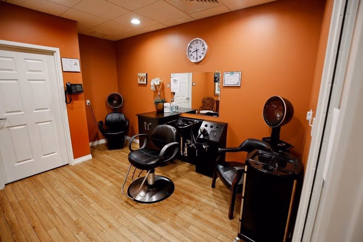 Interior view of a hair salon area with orange walls and wooden flooring. The room contains two black salon chairs, a hair washing station, a mirror, a clock on the wall, and some decorative items including a small plant and a butterfly picture.