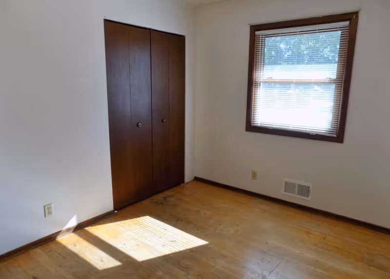 Empty room with wooden floor, a window with blinds on the right wall, and a closed wooden closet door on the left wall. Sunlight is casting a shadow on the floor.