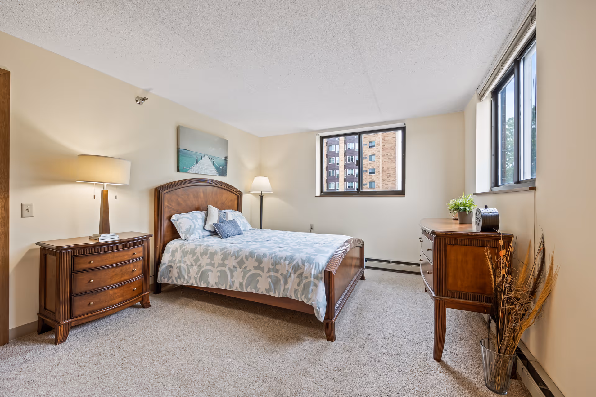 A bright bedroom with a wooden bed frame and a patterned bedspread, two wooden dressers, a table lamp on one dresser, a floor lamp in the corner, a framed picture above the bed, and two windows showing an outside view of a brick building.
