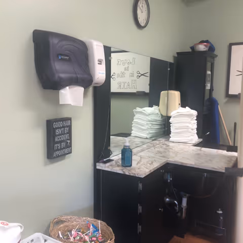 Interior view of a hair salon station with a wall-mounted paper towel dispenser, a sign that reads 'GOOD HAIR ISN'T BY ACCIDENT, IT'S BY APPOINTMENT,' a large mirror, a countertop with neatly stacked white towels, a bottle of hand sanitizer, and a cabinet below the counter with visible plumbing. A clock is mounted on the wall above the mirror.