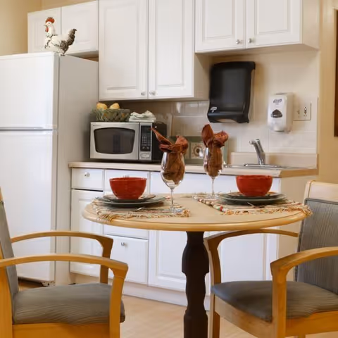 Small kitchenette with a round table set for two in front of white cabinets, microwave, sink, and refrigerator.
