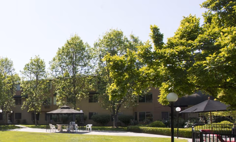 Outdoor view of Bethel Retirement Community showing a grassy area with trees, a paved walkway, and a gazebo with chairs underneath. The building is visible in the background with multiple windows.