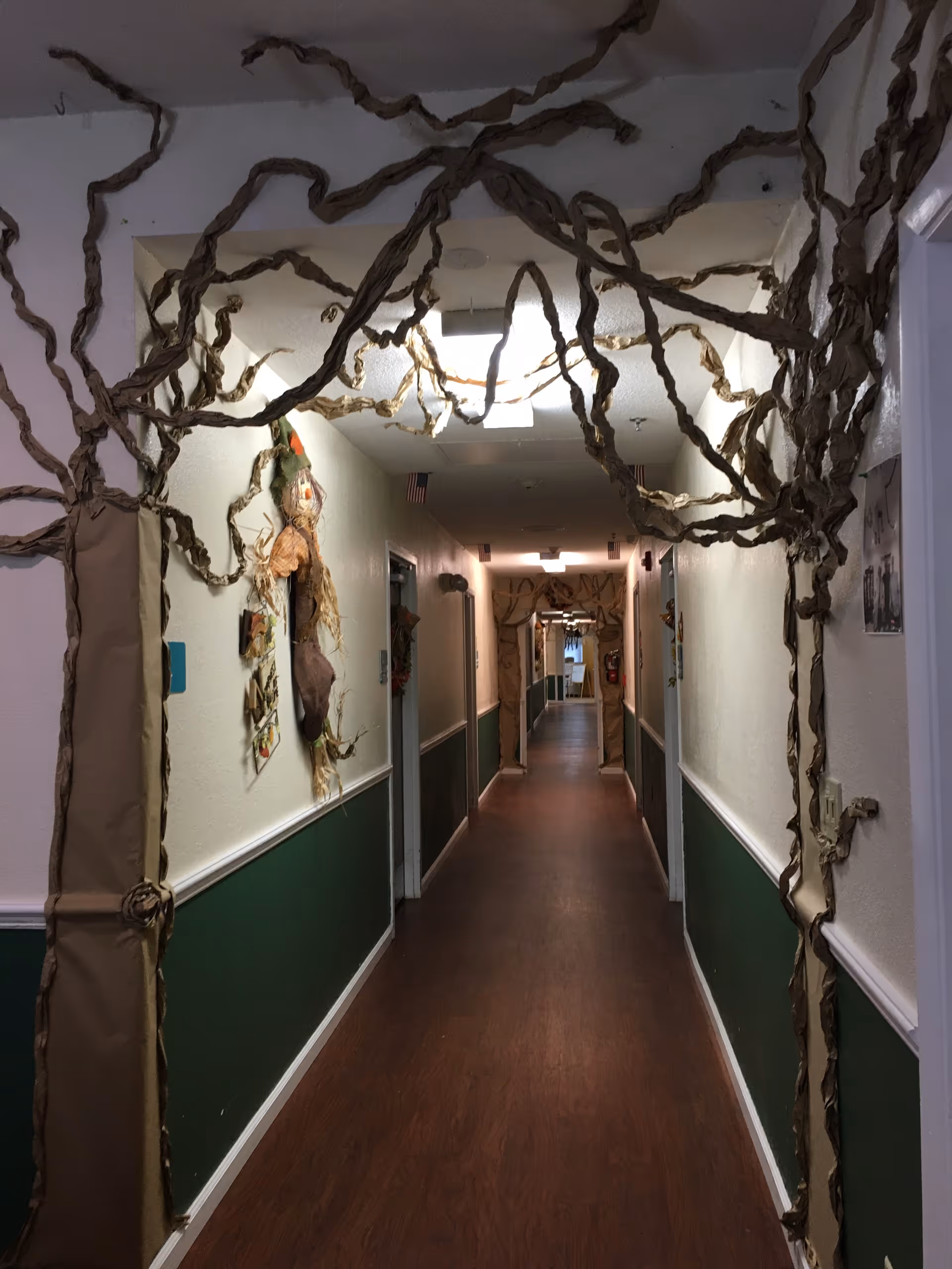 A long indoor hallway in Somerset Assisted Living & Memory Care decorated with twisted brown paper resembling tree branches arching over the entrance and along the walls. The hallway has green and white walls, wooden flooring, and several doors along each side. There are some wall decorations including a scarecrow figure and small American flags hanging from the ceiling.