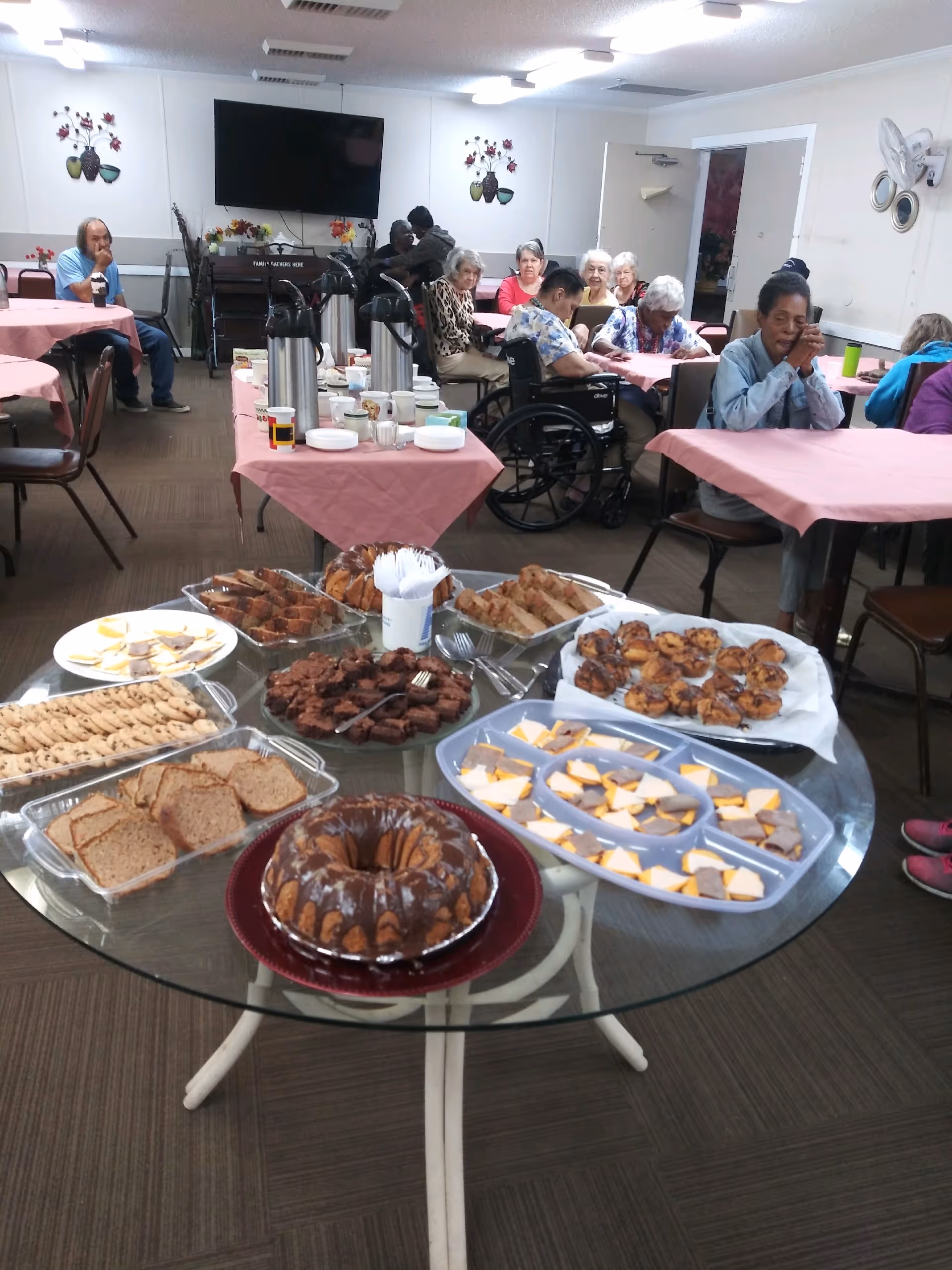 A group of elderly people sitting at tables covered with pink tablecloths in a common dining area. In the foreground, a round glass table displays an assortment of baked goods and snacks including cookies, brownies, cheese and crackers, and a bundt cake. The room has a television mounted on the wall and decorative wall hangings.