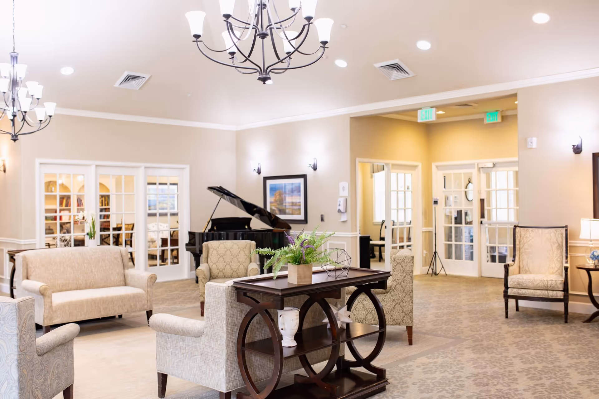 A bright and spacious living room area in a senior living facility featuring comfortable upholstered chairs and sofas arranged around a dark wooden table with decorative plants. A grand piano is visible in the background near glass-paneled double doors, with soft lighting from chandeliers and wall sconces illuminating the beige walls and patterned carpet.
