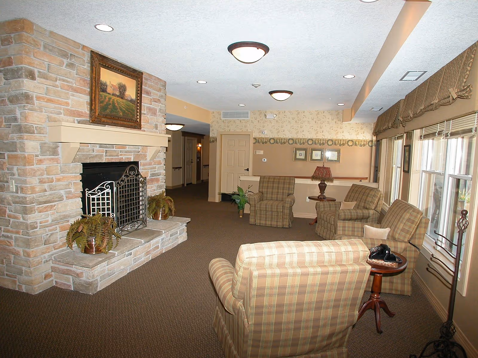 A cozy living room area in a senior living facility featuring a stone fireplace with a painting above it, several plaid upholstered armchairs arranged around small wooden tables, potted plants, and large windows with valances allowing natural light to fill the space.