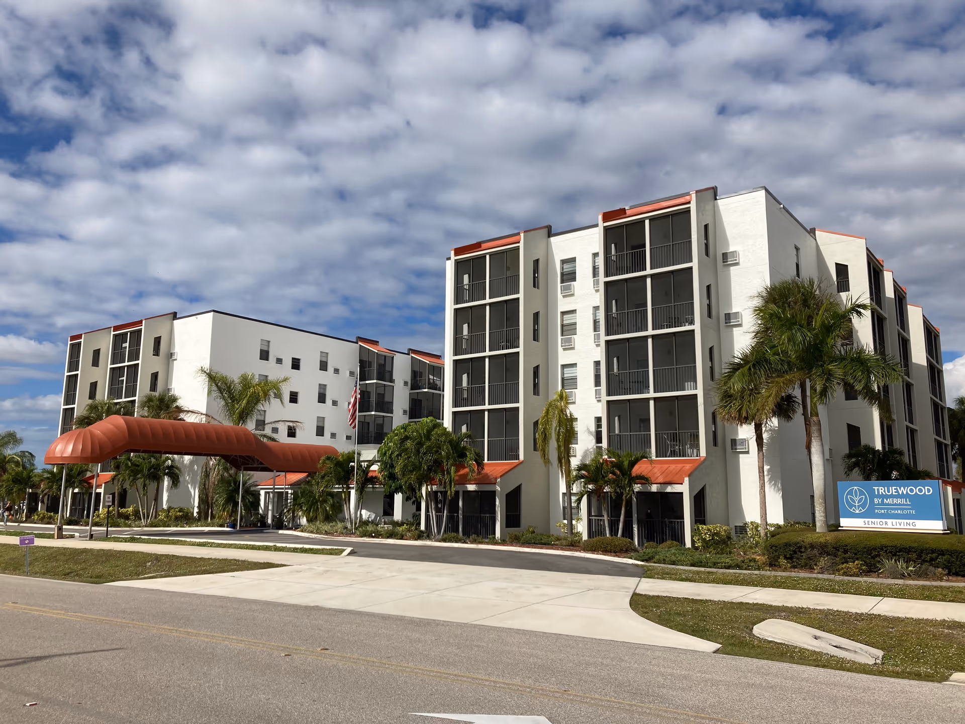 Exterior view of a multi-story senior living facility with white walls and red awnings over entrances and balconies. Palm trees and landscaping surround the building under a partly cloudy sky. A sign in front reads 'Truewood by Merrill Port Charlotte Senior Living.'