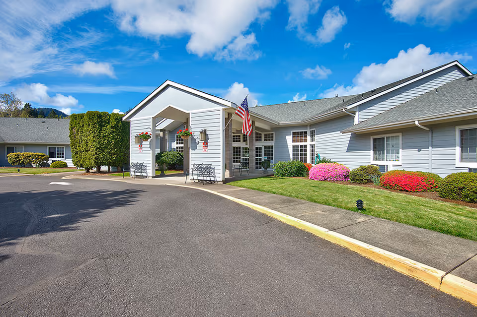 Exterior view of a single-story assisted living facility building with a covered entrance, American flag, well-maintained landscaping including green bushes and colorful flowers, and a clear blue sky with some clouds.