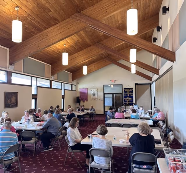 A large room with a high wooden ceiling and hanging lights, filled with elderly people sitting at tables engaged in various activities. The room has large windows on one side and banners on the far wall.