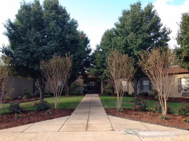 Front entrance of a single-story nursing center with a straight walkway flanked by trees and landscaped beds.