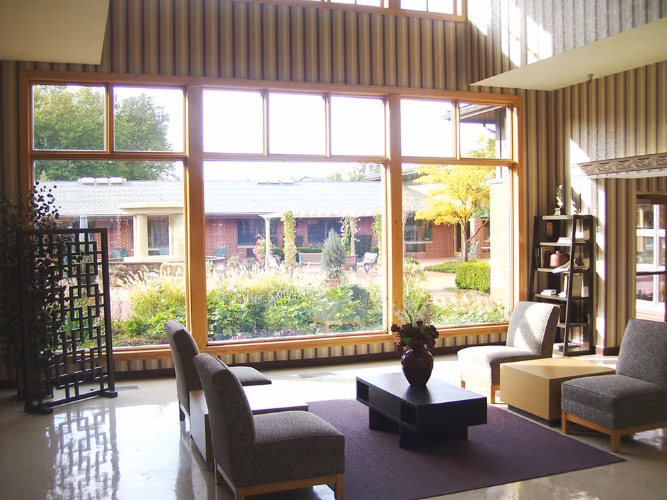 A bright and spacious living room area with large windows overlooking a garden courtyard. The room features four cushioned chairs arranged around a low black coffee table with a vase of flowers on it. There is a purple rug under the table and chairs, a tall black shelving unit with decorative items, and a folding screen near the window. The walls have vertical striped wallpaper and the floor is polished.