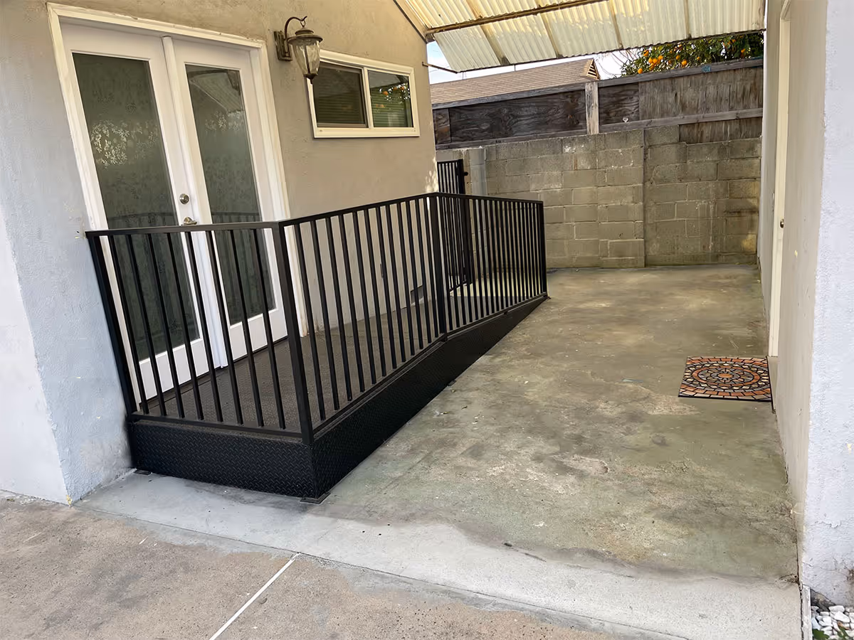 Covered outdoor concrete patio with a black metal wheelchair ramp and railing leading to white French doors and a cinderblock wall.