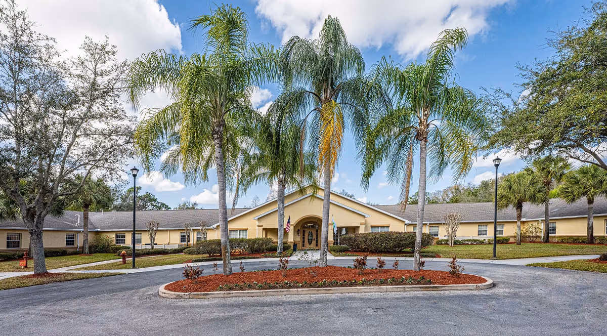Front exterior view of a single-story building with a beige facade and a peaked roof, surrounded by palm trees and other greenery under a partly cloudy sky. There is a circular driveway with a landscaped island in the center featuring palm trees and small plants.