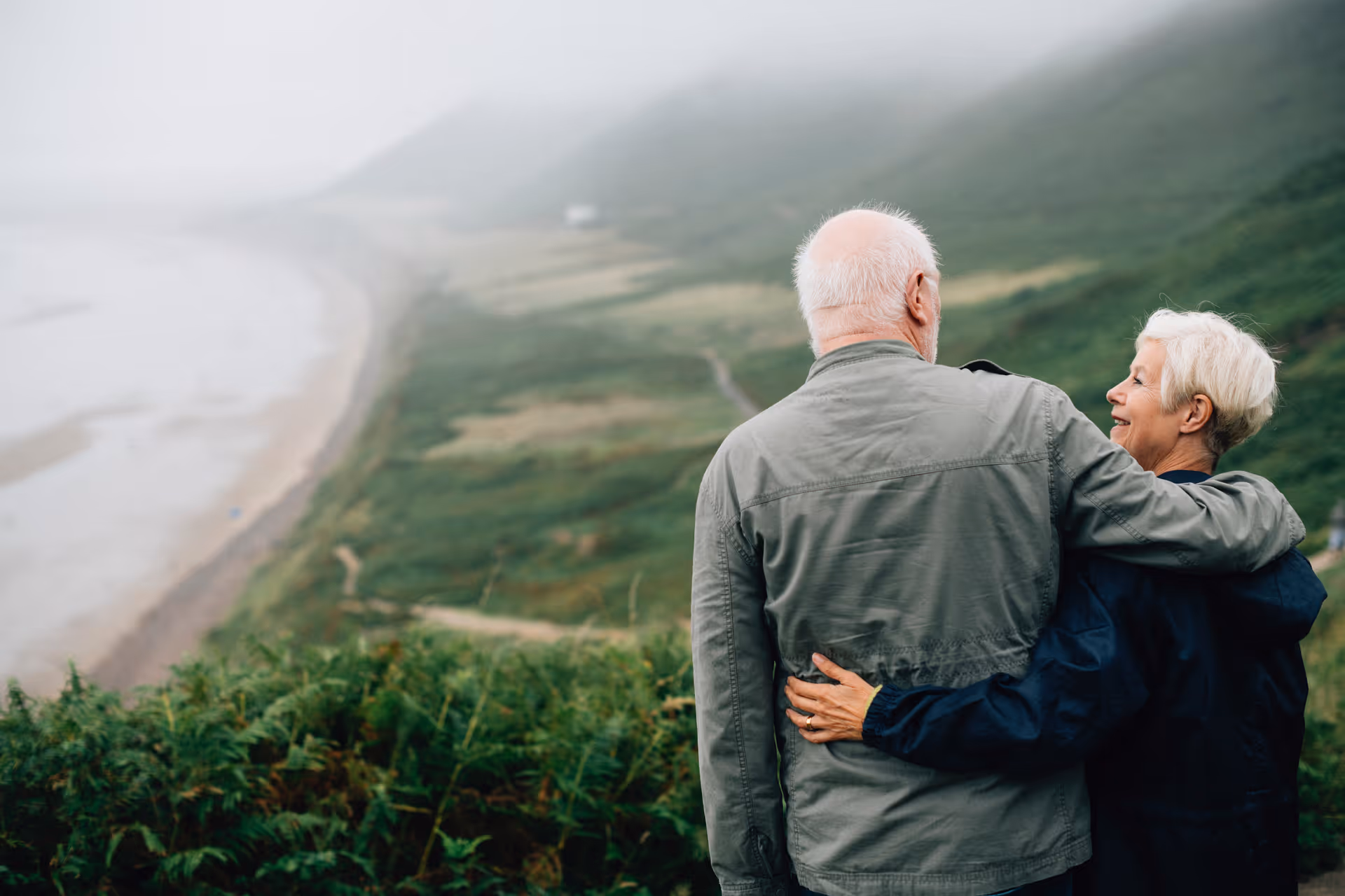 An elderly couple standing outdoors on a grassy hillside overlooking a misty coastline. The man has his arm around the woman, and they are both looking out towards the sea and landscape.
