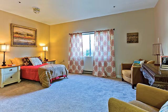 A cozy bedroom with a bed covered in red and beige bedding, two bedside tables with lamps, a window with patterned curtains, a beige carpet, an armchair with a decorative pillow, and a wooden desk with books and a lamp.