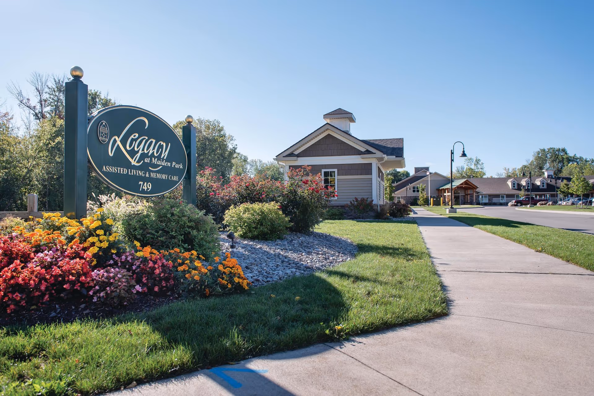 Outdoor view of Legacy at Maiden Park assisted living and memory care facility sign surrounded by colorful flowers and greenery, with a sidewalk and buildings in the background under a clear blue sky.