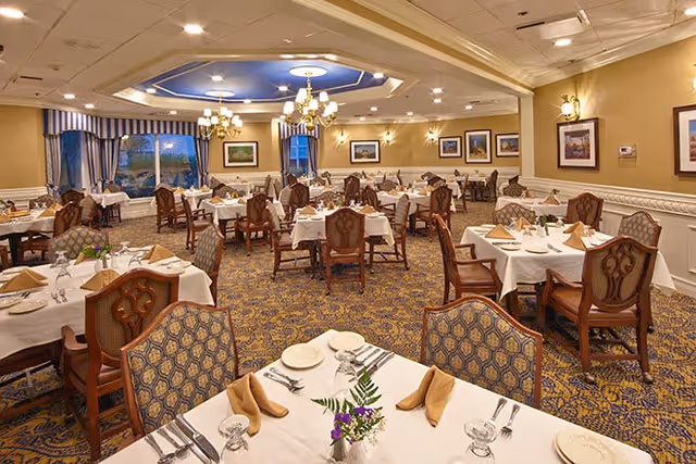 A spacious and elegantly decorated dining room with multiple tables covered in white tablecloths, each set with plates, glasses, silverware, and folded beige napkins. The room features patterned carpet, wooden chairs with upholstered seats and backs, chandeliers hanging from the ceiling, framed artwork on the walls, and large windows with blue and white curtains.
