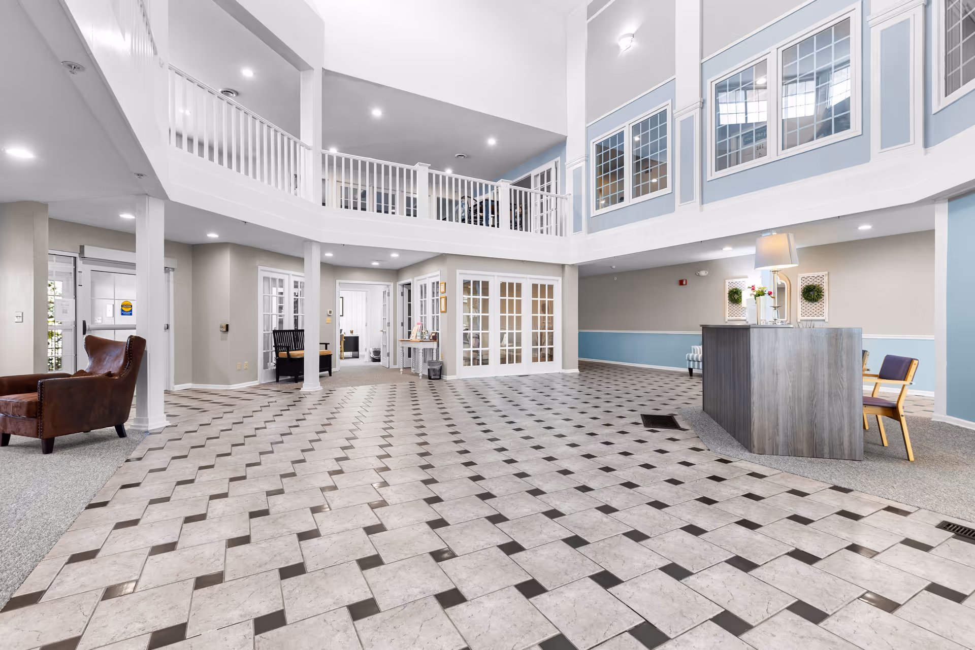 Spacious and bright lobby area of an assisted living facility with a high ceiling, tiled floor, a reception desk with a lamp and flowers, seating areas with chairs, and a second-floor balcony overlooking the space.