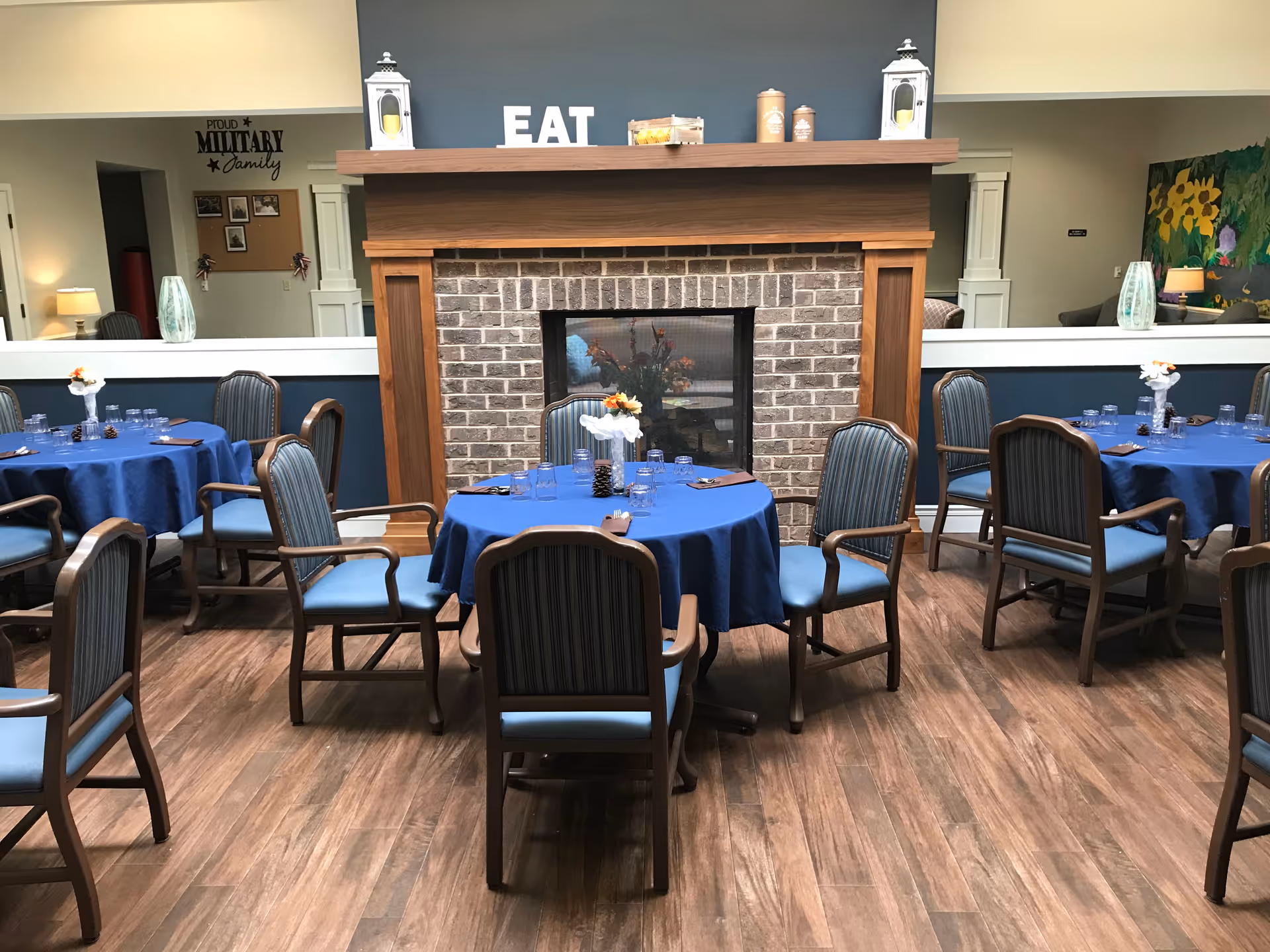 Dining area with round tables covered in blue tablecloths, each set with glasses, napkins, and small floral centerpieces. The room features wooden chairs with blue cushions, a wooden floor, and a brick fireplace with a wooden mantle. On the mantle are decorative items including two white lanterns and letters spelling 'EAT'. The background includes a wall with a 'Proud Military Family' sign and framed photos, as well as a colorful sunflower painting on the right side.