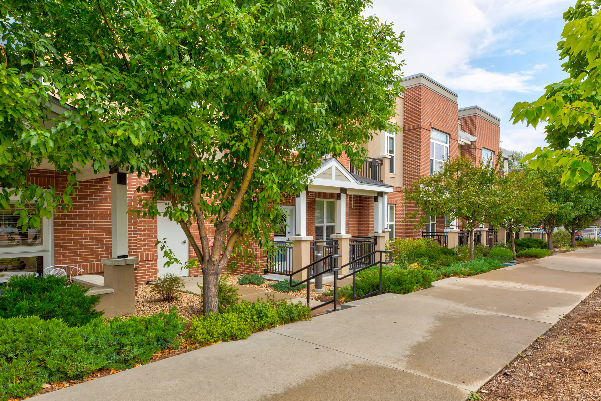 Front exterior of a brick senior living building with trees, landscaped shrubs, a sidewalk, and accessible entry ramps.
