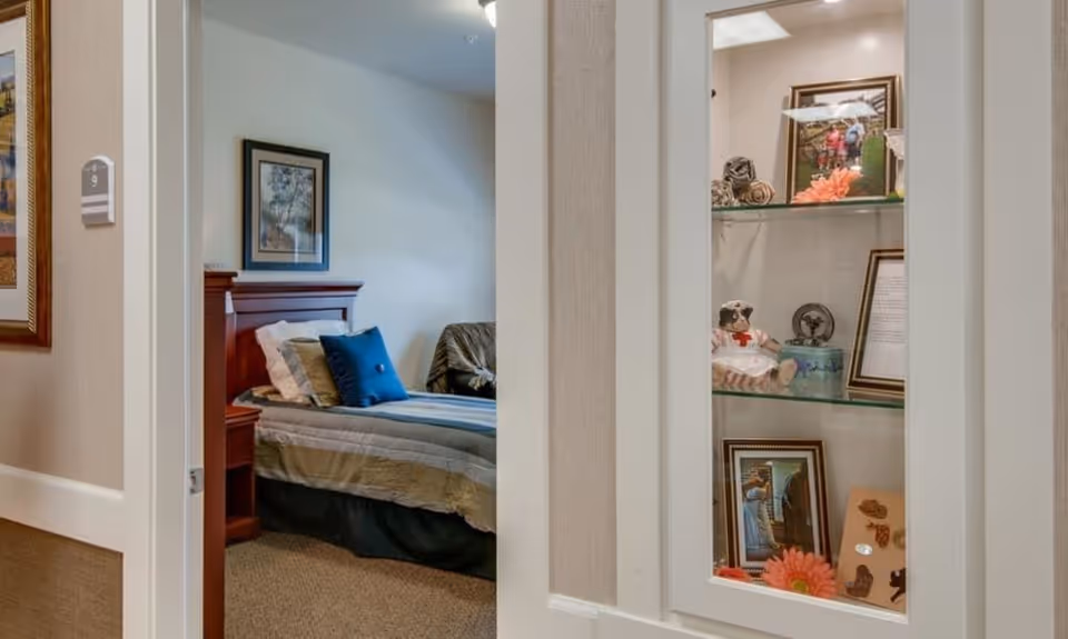 Interior view through a doorway into a furnished bedroom with a made bed, decorative pillows, and a glass-front display cabinet holding framed photos and figurines.