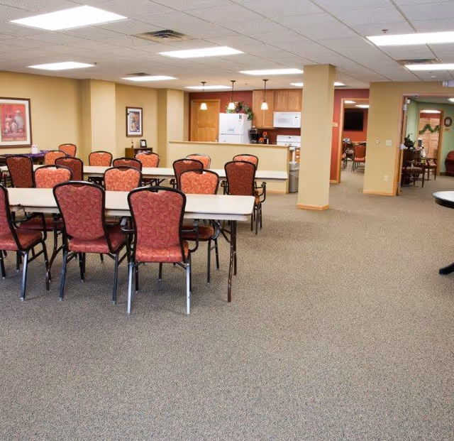 A spacious dining area with multiple rectangular tables and red cushioned chairs arranged neatly. In the background, there is a small kitchen area with wooden cabinets, a white refrigerator, microwave, and coffee maker. The room has beige walls, carpeted floors, and fluorescent ceiling lights. There are framed pictures on the walls and an open doorway leading to another room with more seating visible.