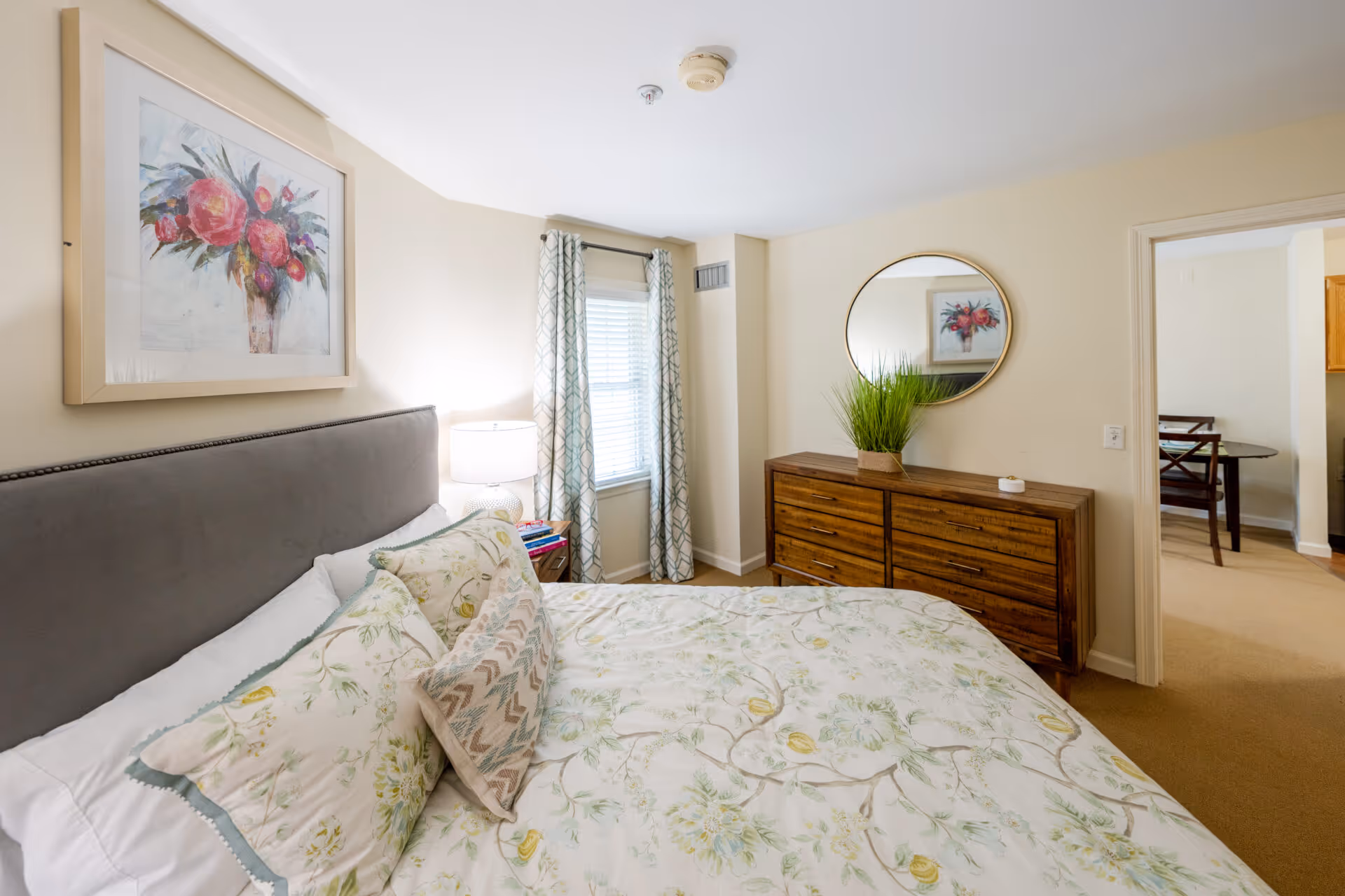 A cozy bedroom with a bed featuring floral bedding and multiple pillows. There is a nightstand with a lamp and books next to a window with patterned curtains. On the opposite wall, there is a wooden dresser with a round mirror above it and a green plant on top. The room has beige walls and carpeted floor, with a doorway leading to another room.