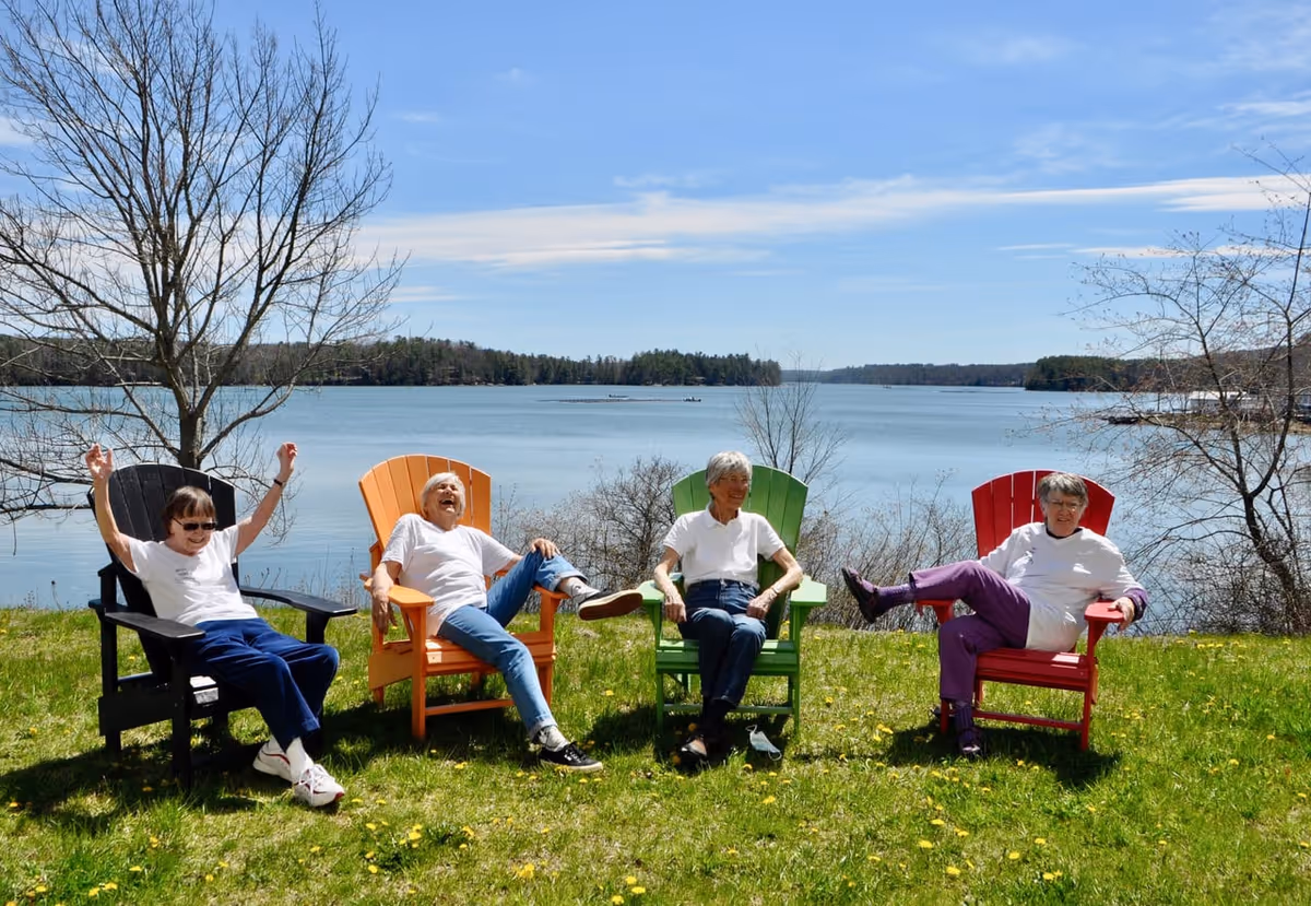 Four elderly women sitting and relaxing on colorful Adirondack chairs on a grassy area near a large lake with trees and a clear blue sky in the background.