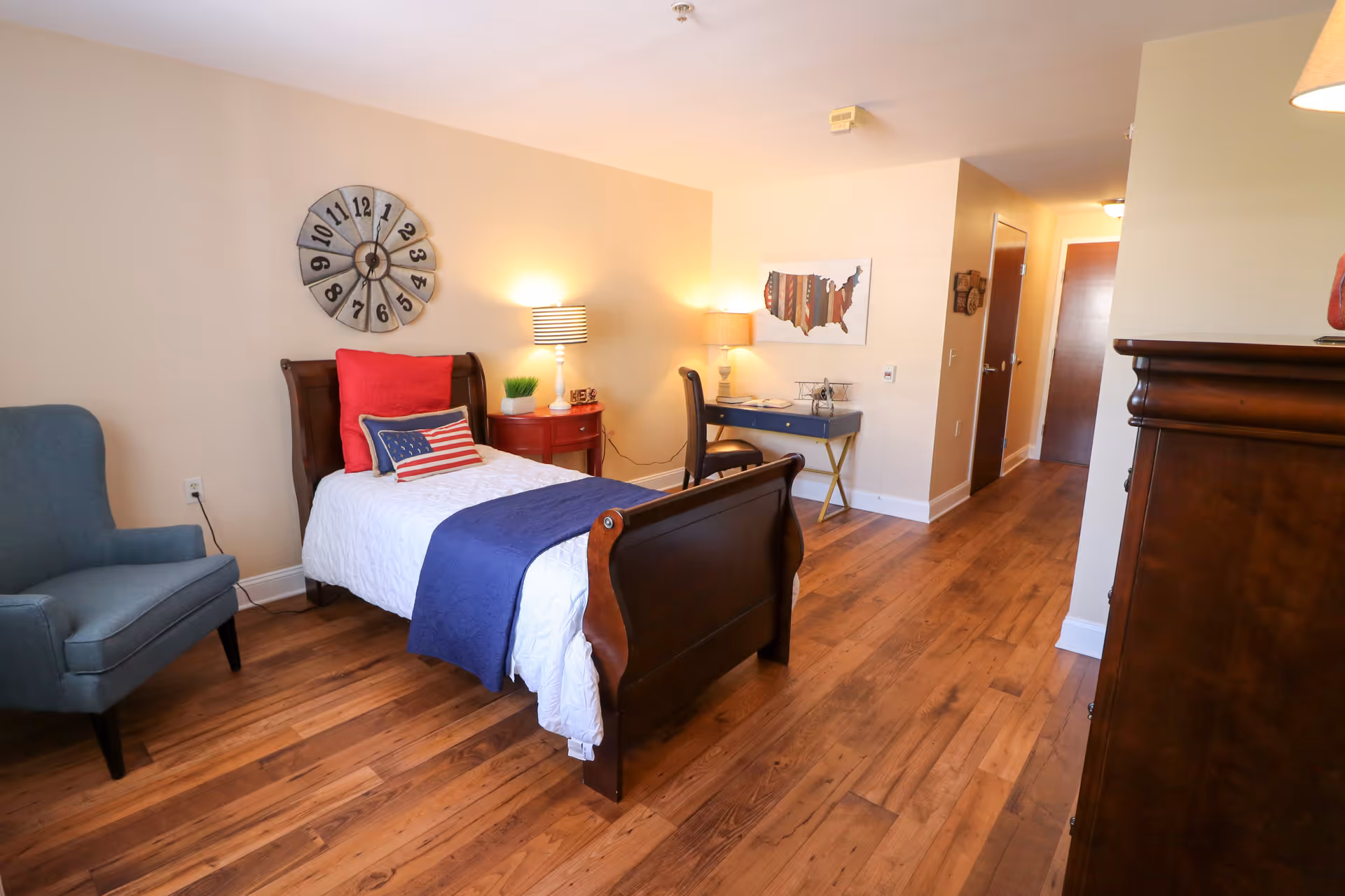 A cozy bedroom with a single wooden bed featuring white bedding, a blue throw blanket, and patriotic pillows. There is a blue armchair on the left, a red nightstand with a lamp, and a desk with a chair and lamp against the far wall. The room has wooden flooring, beige walls, a large decorative clock above the bed, and a framed map of the United States on the wall near the desk.