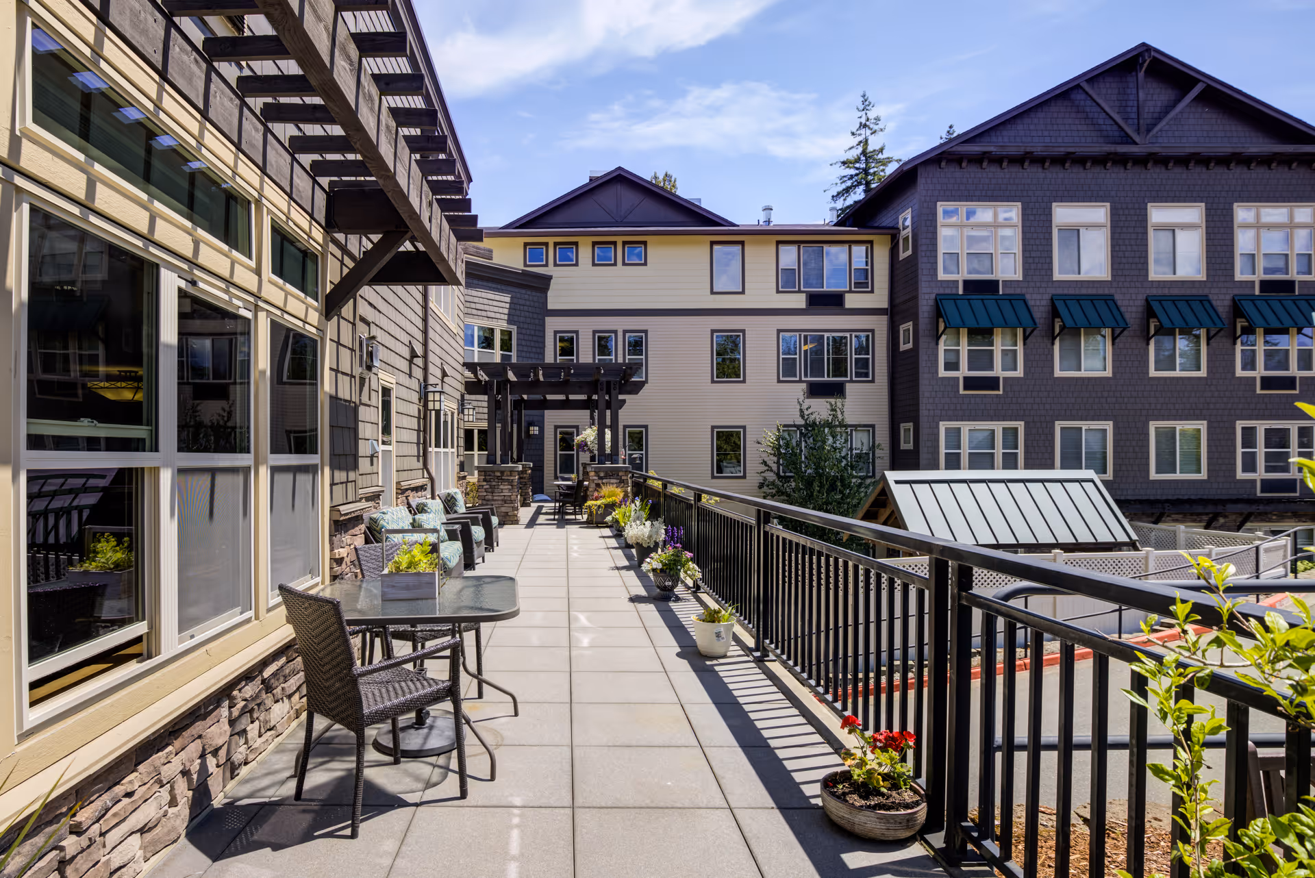 Elevated outdoor patio walkway with tables, chairs, planters and a railing between multi-story buildings.