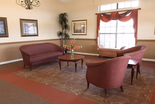 A cozy sitting area with a maroon sofa, two matching armchairs, and a wooden coffee table with a vase of flowers. The room has beige walls with a brown chair rail, framed artwork, a tall potted plant, a large window with red drapes, and a chandelier overhead.